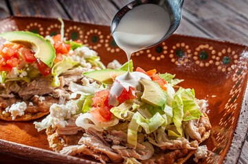 Traditional Mexican food tostadas on a clay or mud plate over a rusic table with chicken or carnitas, lettuce, tomato, onion, fried beans, spicy red salsa and cream. spoon putting cream on top