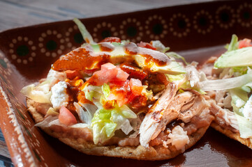 Traditional Mexican food tostadas on a clay or mud plate over a rusic table with chicken or carnitas, lettuce, tomato, onion, fried beans, spicy red salsa and cream. Macro photography