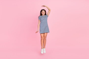 Photo of cute charming girl wear striped dress jumping high measuring height isolated pink color...