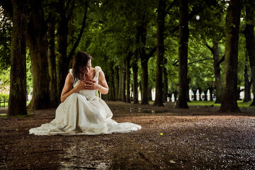 bride in white dress after rain completely wet on the ground on her knees between trees on a long...