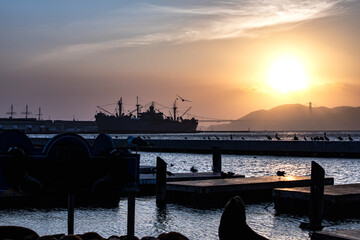 Bright, colorful explosion of twilight sun spreading over a pier in San Francisco with golden orange and yellow rays with the silhouette of a large naval ship and distant Golden Gate Bridge.