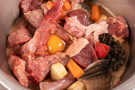 Closeup metal pet bowl with chicken necks, beef meat and bones, paunch, raw egg and carrot pieces. Natural organic daily meal ration preparation for dogs concept. Copy space.