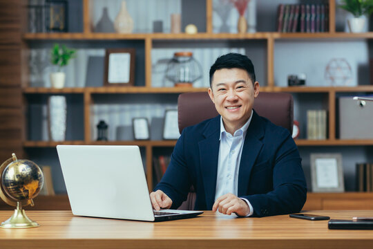 Portrait of a successful Asian businessman, man working in the office sitting at the table, looking at the camera and smiling, happy banker with laptop