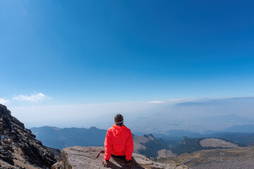 A young cheerful girl in a helmet, sits on the top of the mountain higher than clouds