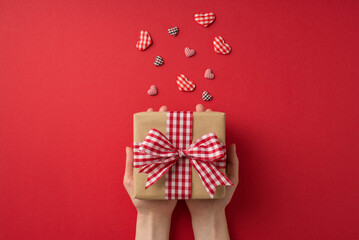 First person top view photo of st valentine's day decorations girl's hands giving craft paper giftbox with checkered ribbon bow and hearts on isolated red background