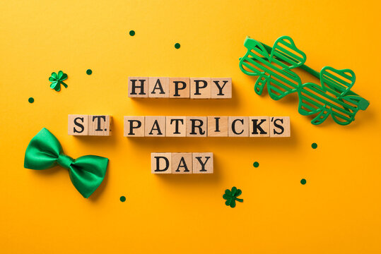 Top view photo of wooden cubes labeled happy st patricks day shamrock shaped party glasses green bow-tie and clover shaped confetti on isolated yellow background