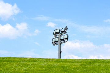 Electric spotlight on a metal pole during a sunny day on a green lawn. White clouds in the blue sky in the background