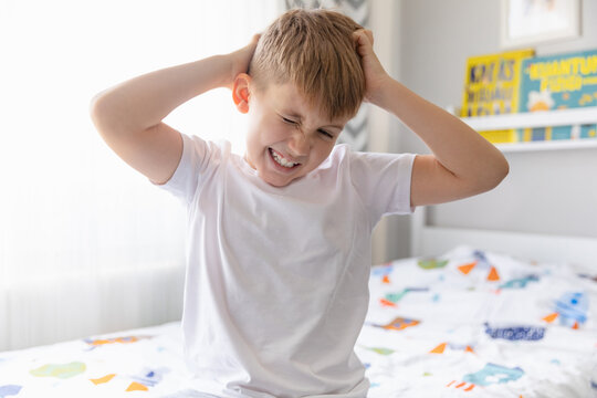 Child boy in white t-shirt sitting on the bed in sunny white room, scratching his head. Cooties, skin diseases concept.