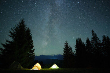 Bright illuminated tourist tents glowing on camping site in dark mountains under night sky with sparkling stars. Active lifestyle concept - obrazy, fototapety, plakaty