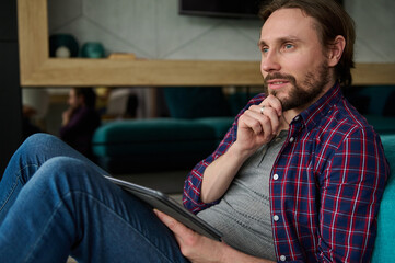 Pensive handsome Caucasian man in casual denim and checkered shirt holding a digital tablet and looking thoughtfully away, sitting at home environment