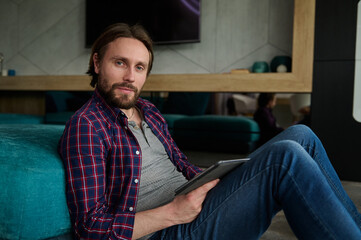 Fototapeta premium Confident handsome bearded Caucasian man in casual jeans and plaid shirt holding digital tablet and looking at camera, sitting on floor with back against sofa while working from home