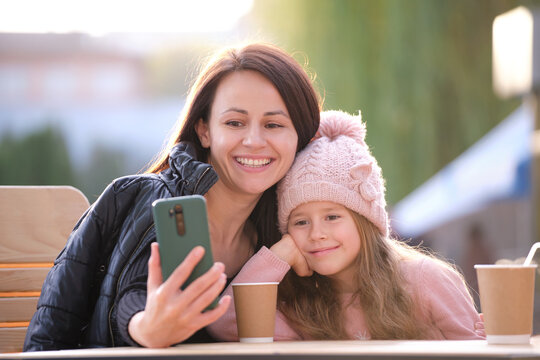 Young mother and her child daughter taking picture with phone selfie camera sitting at street cafe with hot drinks on sunny fall day. Social media presence in everyday life concept