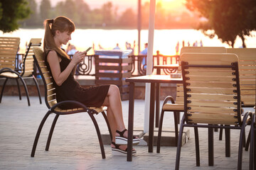 Young woman sitting at street cafe table browsing her cellphone outdoors on warm summer evening. Communication and mobile connection concept