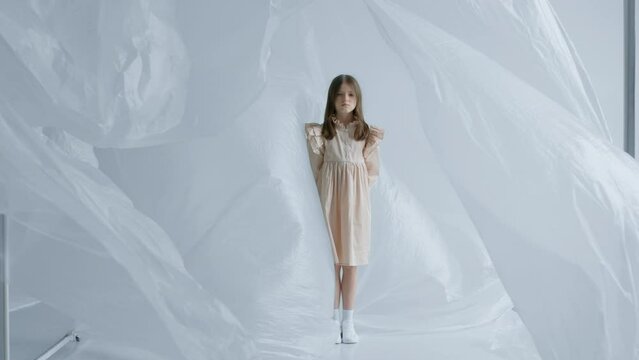 A blonde teen girl stands among the swaying transparent polyethylene in a beige dress with a sad face in abstract space