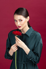 Lovely girl wearing green dress standing isolated over red background, posing with a herbera flower