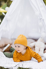 Cute little boy in stylish outfit lying on soft pillow inside colorful wigman among green nature. Portrait of pretty baby posing on fresh air.