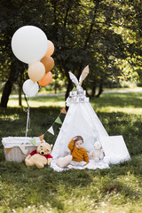 Adorable caucasian boy sitting in colorful wigwam among green nature. Cute child with balloons and toys playing outdoors. Funny time spending. © Sofiia Shunkina