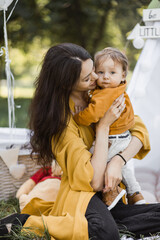 Positive caucasian mother with dark hair sitting with her little son in strong hugs near colorful wigwam. Family of two enjoying summer camping on fresh air.