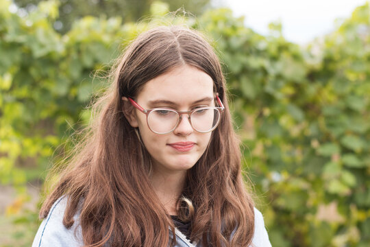 A beautiful girl with braces on her teeth clenching them looks down. Hipster teenager in glasses with long hair against blurred green foliage