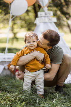 Caring young father hugging and kissing his little son while posing together outdoors. Blur background of colorful balloons and toy teepee tent.