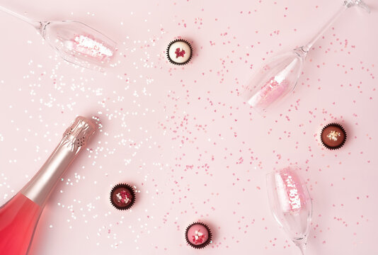 Creative trendy flat lay with pink champagne bottle, flutes and chocolate candy on pink heart shaped confetti background. Flat lay, top view, copy space. Happy Valentines Day greeting card.