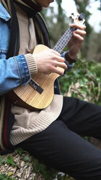 An adult smiling man plays the ukulele in the woods by the river. A strong guy sits and plays a small guitar, against the background of the forest. A happy person plays a musical instrument.