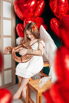 a 5-year-old girl holding a harp for Valentine's Day, in love with red balloons in a white dress and a gold laurel wreath