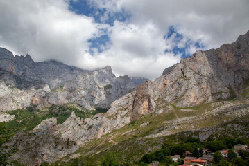 View of the rocky peaks of the Pyrenees, covered with low clouds. Green trees and roofs of houses in the foreground.