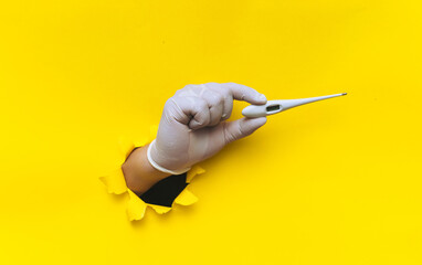 A right hand in a white medical latex glove is pushed through a torn hole in bright yellow paper and holds an electronic thermometer. The concept of diagnosing colds, covid-19, coronavirus. Copy space