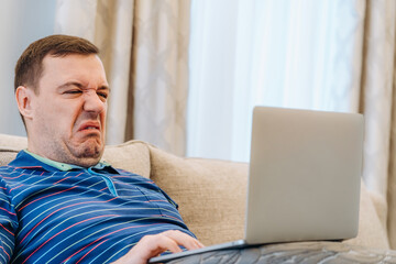 Annoyed millennial man with disgust confused looking at laptop screen sitting on sofa at living room. cringe guy looking at computer with squeamishness, reacting on watching disgusting video at home - obrazy, fototapety, plakaty