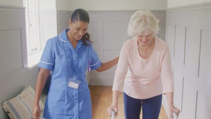 Senior woman at home using walking frame being helped by female care worker in uniform - shot in slow motion