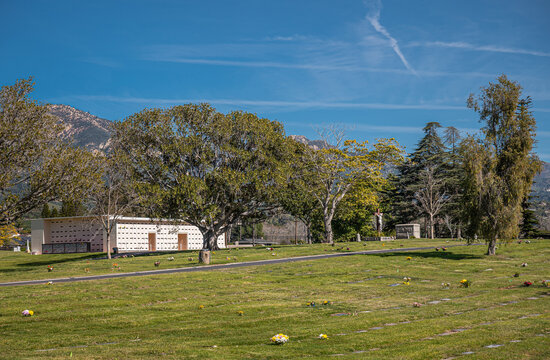 Santa Barbara, California, USA - February 8, 2022: Calvary Cemetery. Wide landscape of burial lawn with smaller wall burial building between lotos of trees under blue sky and Santy Ynes mountains.
