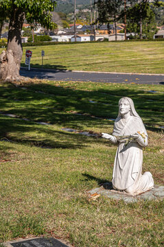 Santa Barbara, California, USA - February 8, 2022: Calvary Cemetery. Closeup of  Bernadette of Lourdes statue on green burial lawn. Faded housing buildings in back.