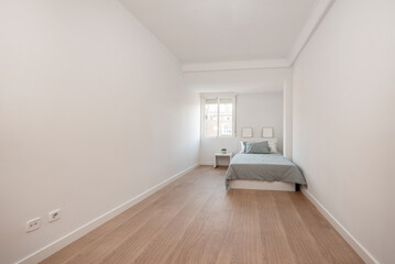 Long bedroom with bed by the window with wooden floorboards and plain white walls