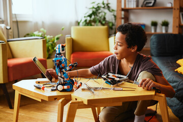 A smart African-American boy holding soldering iron and touching tablet while making a robot at home. Education in electronics.