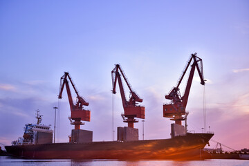 In the setting sun, the crane at the coastal wharf is lifting goods for the cargo ship, with double...