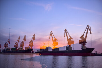 In the setting sun, the crane at the coastal wharf is lifting goods for the cargo ship, with double...