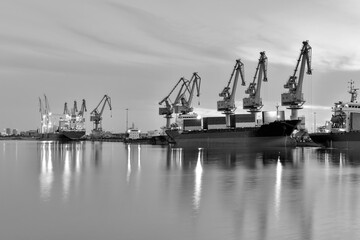Obraz premium In the setting sun, cranes at the coastal wharf are hoisting goods for cargo ships, with monochrome photography effect.
