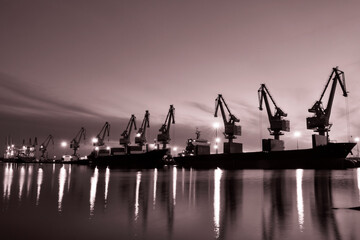 In the setting sun, cranes at the coastal wharf are hoisting goods for cargo ships, with monochrome photography effect.