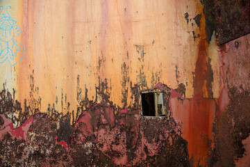 Old rusty metal from a ship that is yellow, gold, red, purple, brown, gray and black. Wet sand and gray barnacles splattered around bottom. Paint is cracked and peeling. Grunge metal background