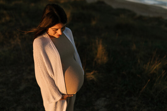 Smiling pregnant woman hugging tummy at sunset
