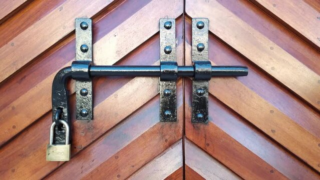 Metal security bar on a wooden double door with padlock. Black steel latch on brown heavy carved gate with fishbone pattern - close up.