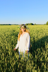 Long haired girl dreesed in white with the frower wreath on the head goes through the corn field in the midsummer day. Blue sky and green meadow