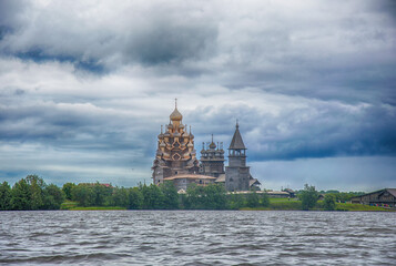 Naklejka premium View of the Kizhi wooden church complex from the water of Onega lake in Karelia Russia. Grey walls of churches and bell tower, dramatic blue sky with grey thunderstorm clouds, green trees, dove water