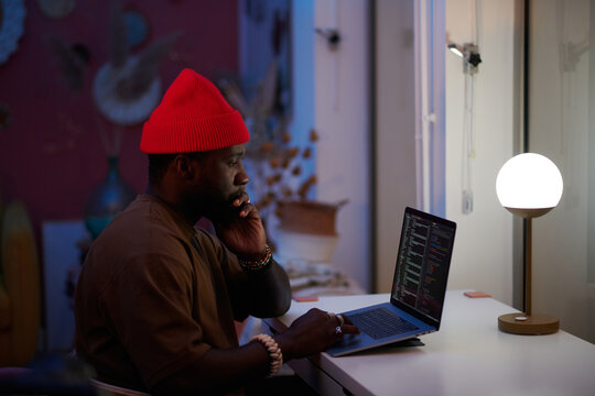 pensive black man with a hat coding on his laptop