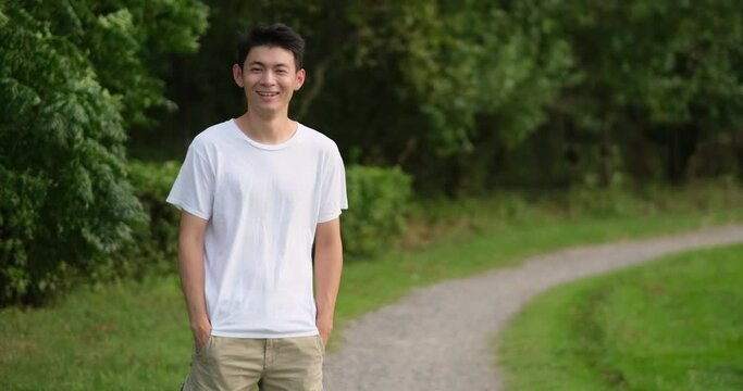Asian young man in white T shirt smiling at camera in green park. Slow motion