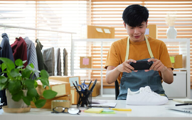 Smiling young asian man online seller using smart phone taking a photo of product for upload to...