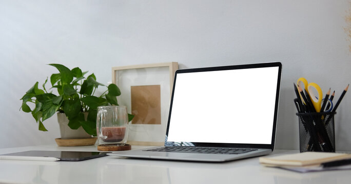 Computer laptop with empty screen, houseplant, picture frame, coffee cup and pencil holder on white table.
