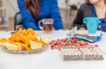 On white table there is dish with fresh slices of apples and tangerines, sweet candies and cookies. Girls are sitting in background.