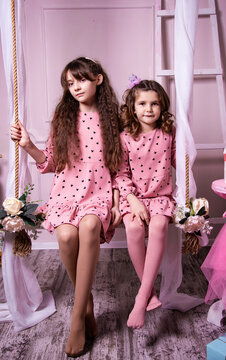 Portrait of two cute girls sitting on a swing in the studio. Children in pink dresses whisper secrets into each other's ears.